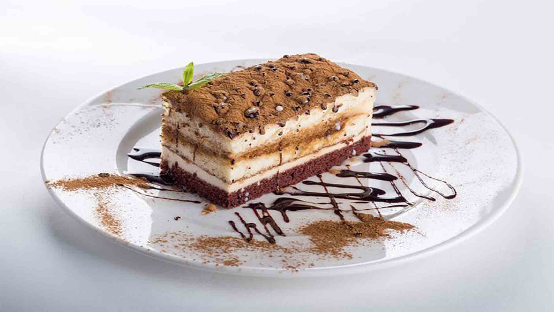 A slice of layered tiramisu cake on a white plate with chocolate drizzle and a dusting of cocoa powder.