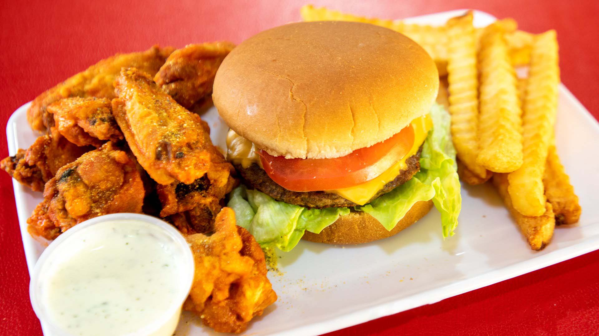 A plate of fast food including a burger, fried chicken wings, crinkle-cut fries, and a side of dip.