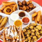 A variety of fried appetizers served with different dipping sauces on a white platter against a red background.
