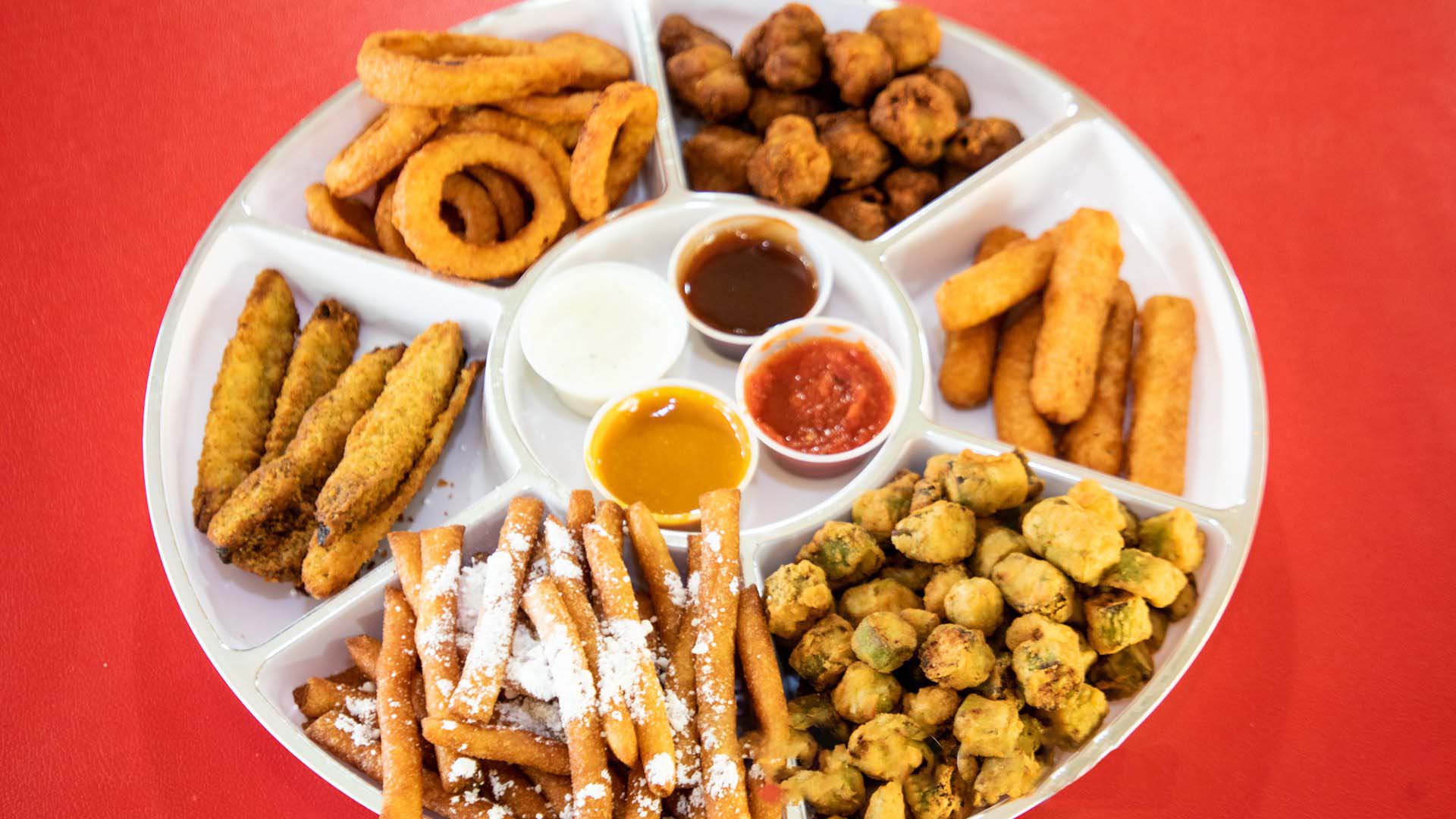 A variety of fried appetizers served with different dipping sauces on a white platter against a red background.