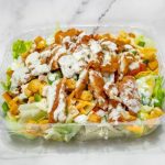 Plastic container with a mixed salad topped with crispy chicken and drizzled with dressing on a marble surface.