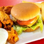 A plate of fast food including a burger, fried chicken wings, crinkle-cut fries, and a side of dip.